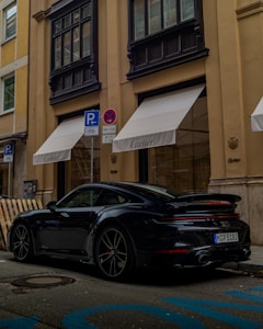 A sleek black sports car is parked on a city street beside a luxury boutique with 'Cartier' branding. The building features large windows with black frames and beige walls. Blue parking markings are visible on the road, along with signage indicating parking regulations.