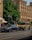 Two luxury vehicles, a sleek gray sedan and a rugged beige SUV, are parked on a city street in front of an ornate historic building with arched windows and detailed stonework. A lush green tree provides shade in the background, while a pedestrian walks nearby.