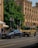 Two luxury vehicles, a sleek gray sedan and a rugged beige SUV, are parked on a city street in front of an ornate historic building with arched windows and detailed stonework. A lush green tree provides shade in the background, while a pedestrian walks nearby.