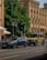 Two luxury vehicles, a sleek gray sedan and a rugged beige SUV, are parked on a city street in front of an ornate historic building with arched windows and detailed stonework. A lush green tree provides shade in the background, while a pedestrian walks nearby.