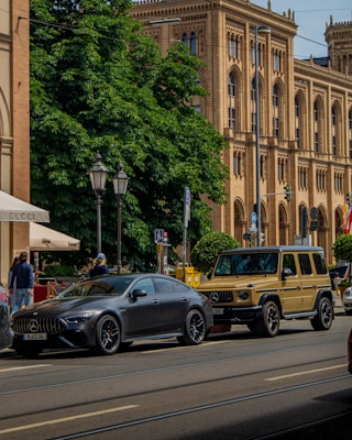 Two luxury vehicles, a sleek gray sedan and a rugged beige SUV, are parked on a city street in front of an ornate historic building with arched windows and detailed stonework. A lush green tree provides shade in the background, while a pedestrian walks nearby.