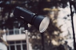 A professional photography light, labeled AD600Pro, mounted on a metal stand. The background is slightly out of focus, showing blurred trees and a white building.