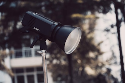 A professional photography light, labeled AD600Pro, mounted on a metal stand. The background is slightly out of focus, showing blurred trees and a white building.