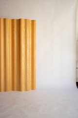 A minimalist interior featuring a vertical wooden panel against a smooth, light-colored backdrop. The panel displays a natural wood grain pattern and is positioned to one side, creating a subtle contrast with the plain surroundings.