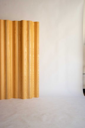 A minimalist interior featuring a vertical wooden panel against a smooth, light-colored backdrop. The panel displays a natural wood grain pattern and is positioned to one side, creating a subtle contrast with the plain surroundings.