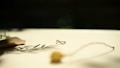 A small, delicate ring with a loop design sits on a light surface. There is a soft, golden lighting effect, creating gentle shadows. A blurred twig and an out-of-focus fabric piece are placed nearby, adding texture to the image.