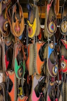 A collection of colorful handmade leather shoes hangs on display. Each shoe features vibrant, unique patterns and is stitched with contrasting thread. The colors range from bright yellow and pink to deep brown and black, creating a lively and artistic arrangement.