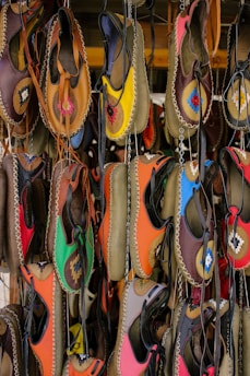 A collection of colorful handmade leather shoes hangs on display. Each shoe features vibrant, unique patterns and is stitched with contrasting thread. The colors range from bright yellow and pink to deep brown and black, creating a lively and artistic arrangement.