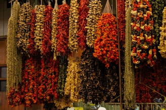 Multiple strands of dried vegetables and herbs, including red peppers, hung from the ceiling in a market setting. The arrangement appears vibrant and rustic, creating a display of texture and color.