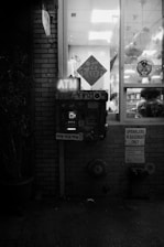 An ATM is situated outside a building with brick walls. The ATM is illuminated by a glowing sign above it. To the right, a glass window displays hanging decorations and a glimpse into a room. Various signs are posted on the wall, including one indicating sprinklers in the basement.