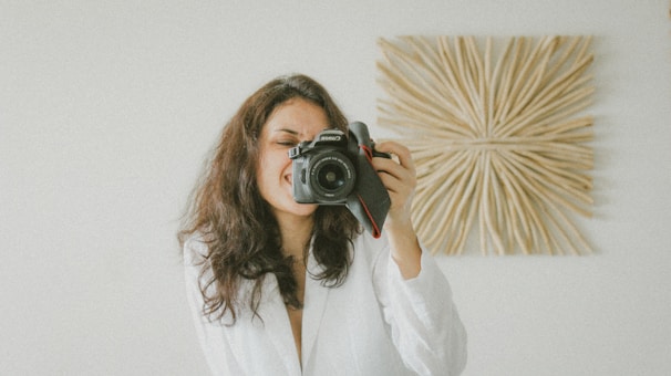 A person with long dark hair is holding a camera up to their face, capturing a photo. They are wearing a white garment and smiling. In the background, there is a decorative wall hanging design featuring a sunburst pattern.