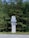 A tall sign with Cyrillic lettering stands in front of a lush background of pine trees. The sign is white with blue elements, featuring vertical lines and decorative structures.