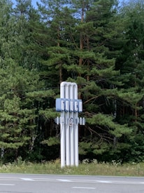 A tall sign with Cyrillic lettering stands in front of a lush background of pine trees. The sign is white with blue elements, featuring vertical lines and decorative structures.