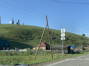 A rural landscape with wooden houses set against a grassy hillside. Utility poles and wires run alongside the road. The sky is clear with a few clouds, and there are several trees on the hilltop.