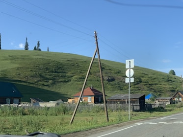 A rural landscape with wooden houses set against a grassy hillside. Utility poles and wires run alongside the road. The sky is clear with a few clouds, and there are several trees on the hilltop.