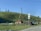 A rural landscape with wooden houses set against a grassy hillside. Utility poles and wires run alongside the road. The sky is clear with a few clouds, and there are several trees on the hilltop.