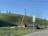 A rural landscape with wooden houses set against a grassy hillside. Utility poles and wires run alongside the road. The sky is clear with a few clouds, and there are several trees on the hilltop.