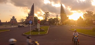 A roundabout at sunset with distinctive split gateway structures typical of Balinese architecture. Several vehicles, including motorcycles and a car, are navigating the road. Trees and a statue are present in the roundabout's center, with a dramatic sky featuring golden sunlight breaking through clouds.