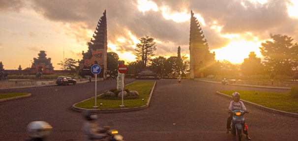 A roundabout at sunset with distinctive split gateway structures typical of Balinese architecture. Several vehicles, including motorcycles and a car, are navigating the road. Trees and a statue are present in the roundabout's center, with a dramatic sky featuring golden sunlight breaking through clouds.