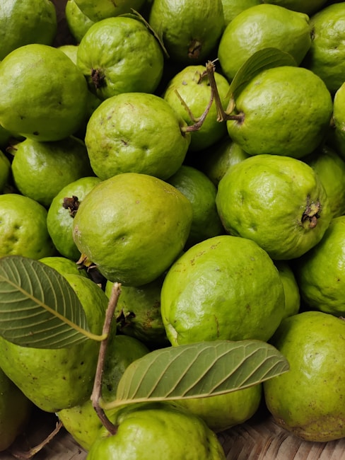 A collection of fresh green guavas piled together, with a couple of leaves interspersed among the fruits. The guavas have a smooth texture and a vibrant green color, indicating ripeness.
