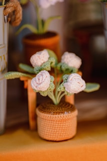 A knitted or crocheted plant with white flowers and green leaves in a woven, brown pot. The plant is crafted with textured yarn, giving it a handmade appearance. It is placed on a surface with warm, earthy tones, and other blurred decorative objects are seen in the background.