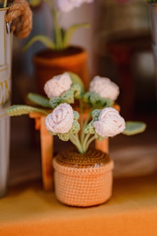 A knitted or crocheted plant with white flowers and green leaves in a woven, brown pot. The plant is crafted with textured yarn, giving it a handmade appearance. It is placed on a surface with warm, earthy tones, and other blurred decorative objects are seen in the background.
