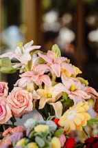 A collection of intricately crafted crochet flowers in various colors including pink, yellow, and white. The flowers resemble lilies, roses, and daisies, and are arranged in a vibrant bouquet. The background is softly blurred, emphasizing the detailed textures of the yarn and the lifelike appearance of the flowers.