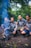 Four men are sitting together in a forested area, wearing matching shirts with 'Rinjani Dawn Adventures' written on them. They are gathered around a small fire with a smoky trail rising upwards. The surrounding environment is lush and green, suggesting a dense woodland setting.