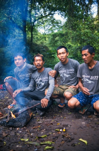 Four men are sitting together in a forested area, wearing matching shirts with 'Rinjani Dawn Adventures' written on them. They are gathered around a small fire with a smoky trail rising upwards. The surrounding environment is lush and green, suggesting a dense woodland setting.