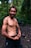 A muscular man smiles confidently in an outdoor forest setting. He is shirtless, demonstrating a strong and well-defined physique. The background is lush with green foliage and the ground is covered with rocks and leaves.