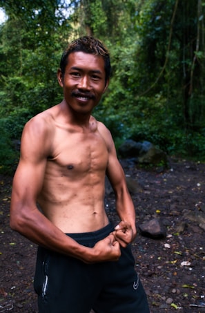 A muscular man smiles confidently in an outdoor forest setting. He is shirtless, demonstrating a strong and well-defined physique. The background is lush with green foliage and the ground is covered with rocks and leaves.