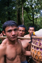 Three shirtless men are standing closely together in a forested environment. They are carrying a woven basket filled with plastic bottles and other items, which is supported on a wooden stick over their shoulders. The background is dense with tall trees and green foliage indicating a lush jungle setting.