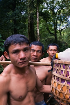 Three shirtless men are standing closely together in a forested environment. They are carrying a woven basket filled with plastic bottles and other items, which is supported on a wooden stick over their shoulders. The background is dense with tall trees and green foliage indicating a lush jungle setting.