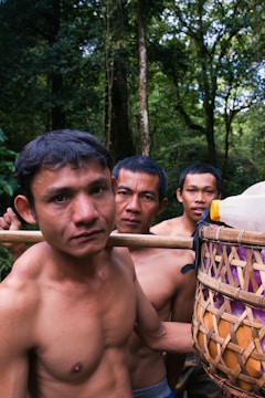 Three shirtless men are standing closely together in a forested environment. They are carrying a woven basket filled with plastic bottles and other items, which is supported on a wooden stick over their shoulders. The background is dense with tall trees and green foliage indicating a lush jungle setting.