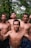 Four shirtless men standing closely together in an outdoor setting with lush green trees in the background. One of the men has a cigarette in his mouth, and they appear to be posing confidently.