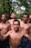 Four shirtless men standing closely together in an outdoor setting with lush green trees in the background. One of the men has a cigarette in his mouth, and they appear to be posing confidently.