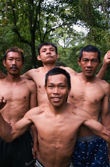 Four shirtless men standing closely together in an outdoor setting with lush green trees in the background. One of the men has a cigarette in his mouth, and they appear to be posing confidently.