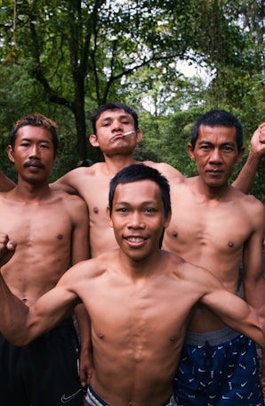 Four shirtless men standing closely together in an outdoor setting with lush green trees in the background. One of the men has a cigarette in his mouth, and they appear to be posing confidently.