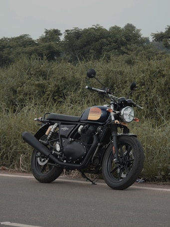 A classic-style motorcycle is parked on a paved road, surrounded by lush green foliage. The motorcycle has a black body with orange accents and features a round headlight, rearview mirrors, and a prominent exhaust pipe.