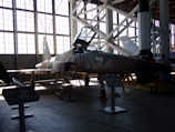 A military jet aircraft displayed inside a large hangar with a high ceiling and large windows. The aircraft, positioned with a ladder next to it, features a camouflage pattern with the number 12 on its side. The hangar is filled with ambient light from the large windows, creating shadows on the ground. Various informational stands and another aircraft or rocket appear in the background.