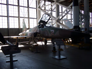 A military jet aircraft displayed inside a large hangar with a high ceiling and large windows. The aircraft, positioned with a ladder next to it, features a camouflage pattern with the number 12 on its side. The hangar is filled with ambient light from the large windows, creating shadows on the ground. Various informational stands and another aircraft or rocket appear in the background.