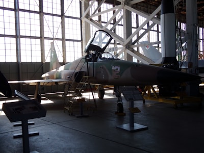 A military jet aircraft displayed inside a large hangar with a high ceiling and large windows. The aircraft, positioned with a ladder next to it, features a camouflage pattern with the number 12 on its side. The hangar is filled with ambient light from the large windows, creating shadows on the ground. Various informational stands and another aircraft or rocket appear in the background.