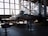 A military jet aircraft displayed inside a large hangar with a high ceiling and large windows. The aircraft, positioned with a ladder next to it, features a camouflage pattern with the number 12 on its side. The hangar is filled with ambient light from the large windows, creating shadows on the ground. Various informational stands and another aircraft or rocket appear in the background.