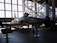 A military jet aircraft displayed inside a large hangar with a high ceiling and large windows. The aircraft, positioned with a ladder next to it, features a camouflage pattern with the number 12 on its side. The hangar is filled with ambient light from the large windows, creating shadows on the ground. Various informational stands and another aircraft or rocket appear in the background.