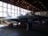 A fighter jet is displayed inside an aviation museum hangar. The aircraft is positioned on the ground with its landing gear deployed. The space is dimly lit, with sunlight filtering through large windows. The scene includes an informational stand in front of the jet.