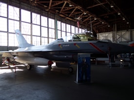 A fighter jet is displayed inside an aviation museum hangar. The aircraft is positioned on the ground with its landing gear deployed. The space is dimly lit, with sunlight filtering through large windows. The scene includes an informational stand in front of the jet.