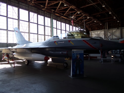 A fighter jet is displayed inside an aviation museum hangar. The aircraft is positioned on the ground with its landing gear deployed. The space is dimly lit, with sunlight filtering through large windows. The scene includes an informational stand in front of the jet.