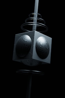 A geometric sculpture features a dark cube with spherical, textured protrusions on each visible face, mounted on a vertical pole. Spiral rings encircle the pole, adding a sense of dynamic movement. The background is black, highlighting the monochromatic design and smooth surfaces of the sculpture.