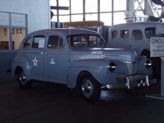 A vintage US Navy car displayed indoors at a museum setting. The car is grey with a small white star and red circle insignia on the door, and has a badge that reads 'US NAVY CUSTOM' on the front. Another vintage vehicle and a sign are visible in the background, along with various garage tools and repair items.