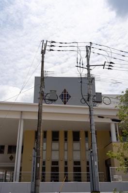 Two utility poles with multiple electrical wires are positioned in front of a modern building with a diamond-shaped window. The building has a yellow and white facade, and the sky is lightly clouded.